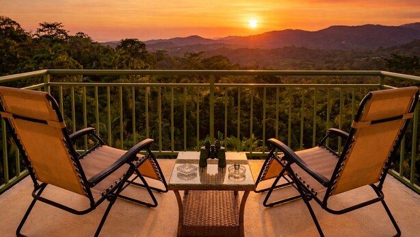Un balcón con dos sillas y una mesa, con vistas a un paisaje montañoso al atardecer.