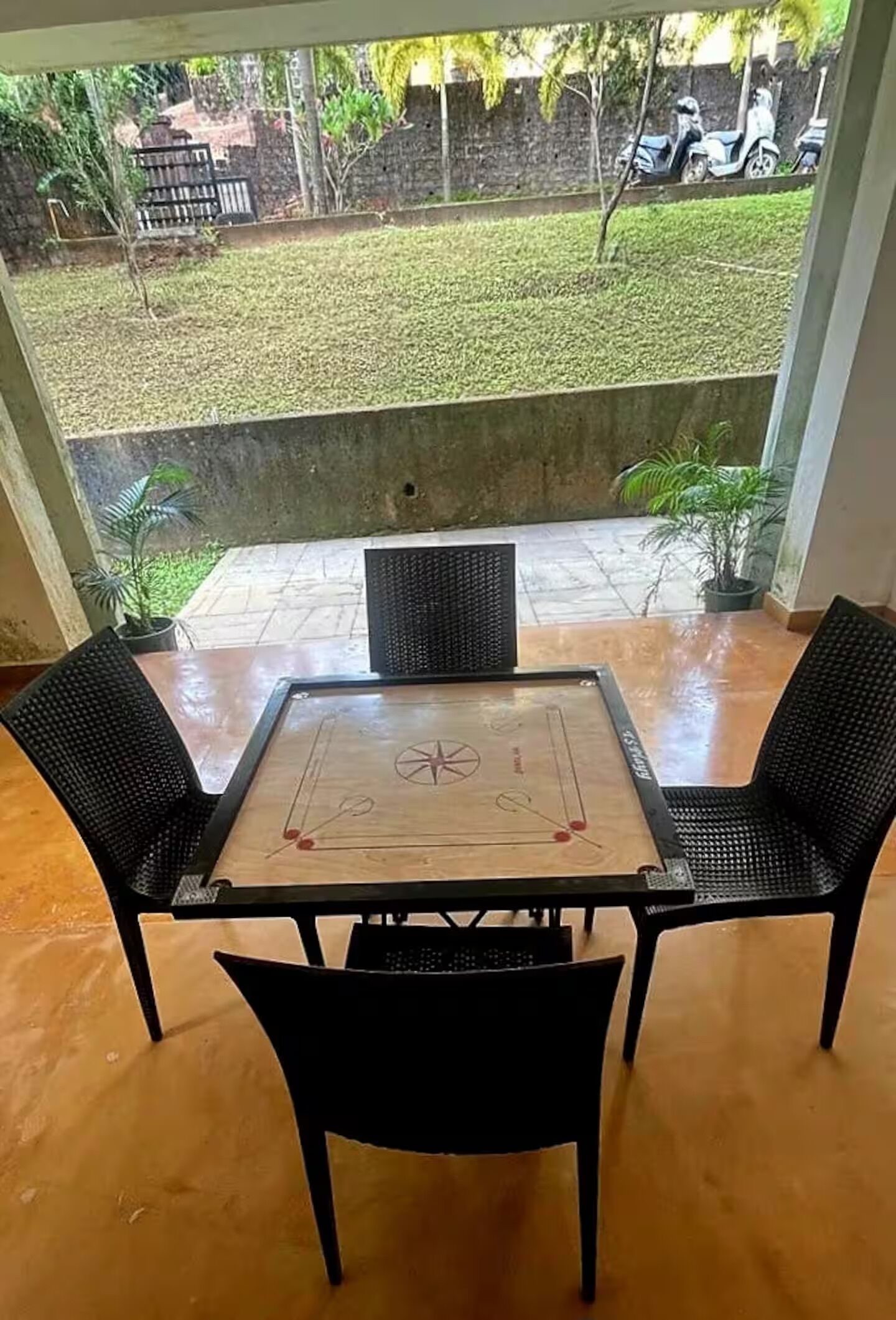 Une pièce avec une table en bois et deux chaises, une vue sur un jardin avec une pelouse et un mur, et un sol en béton.