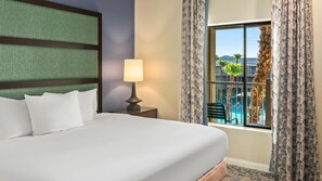 Ein Hotelzimmer mit Bett, Nachttisch mit Lampe, Fenster mit Vorhängen und Blick auf einen Pool.