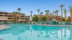 Ein Hotelpoolbereich mit mehreren Liegestühlen und Sonnenschirmen, umgeben von Palmen und einem mehrstöckigen Gebäude.