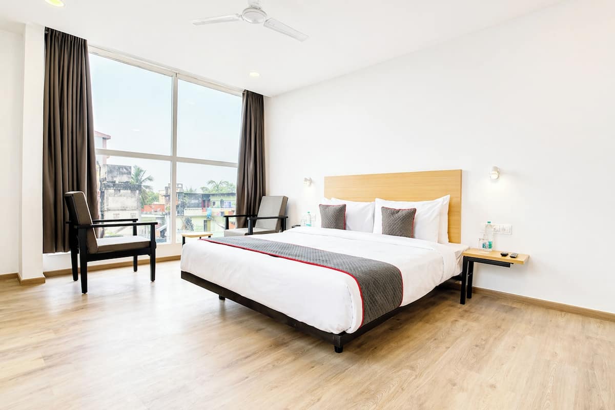 Habitación de hotel con una cama grande, una silla, una mesita de noche y vistas a la ciudad a través de amplios ventanales.