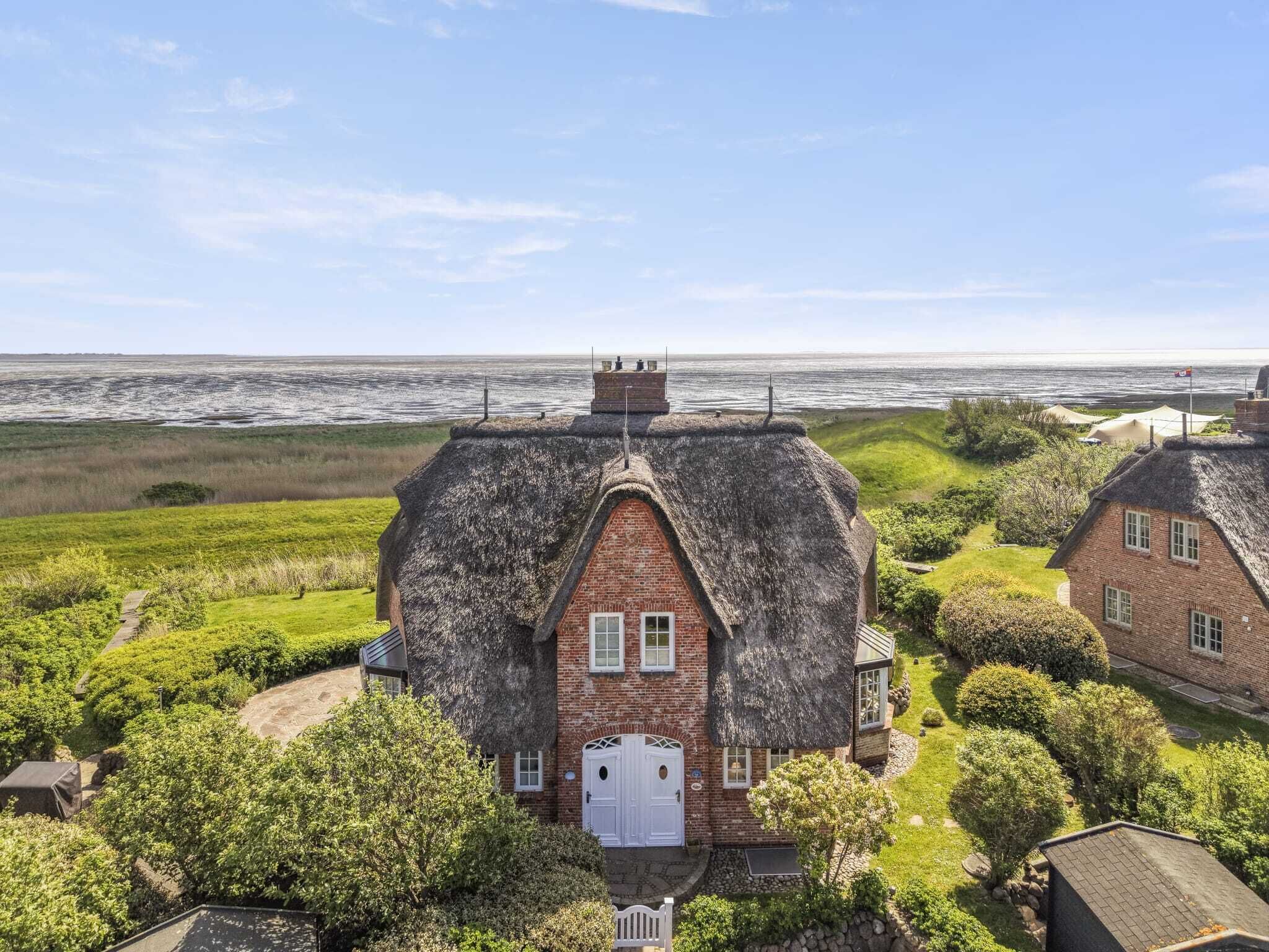 A thatched-roof house with a brick lower section, surrounded by greenery and a view of the sea.