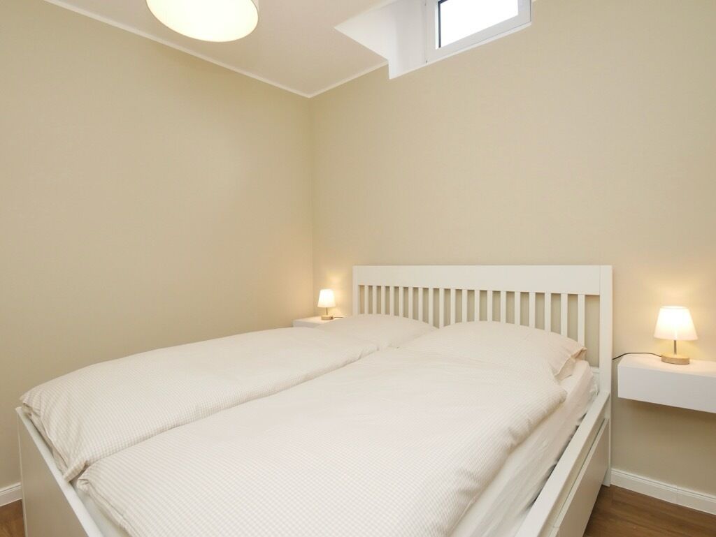 Ein Einzelbett mit weißem Kopfteil, zwei Nachttischlampen und ein kleines Fenster darüber.