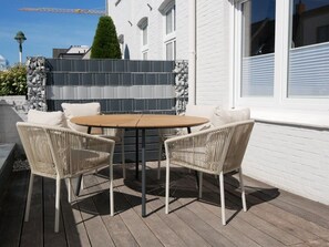 Eine Terrasse mit einem runden Tisch und vier Korbstühlen, eine weiße Ziegelwand und ein Zäunchen aus Fliesen.