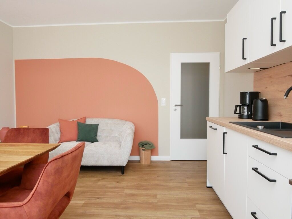 Una cocina moderna con armarios blancos y encimera de madera, un comedor con mesa de madera y sillas rojas, y una sala de estar con sofá y un amplio muro naranja.