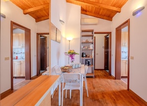 A dining area with a wooden table and chairs, a wall-mounted shelf with kitchen items, and a framed picture above the table. - The Landscape Home (Roma)