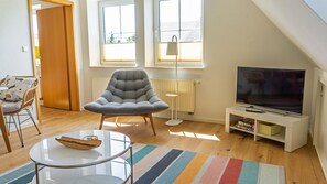 Ein modernes Wohnzimmer mit einem grauen Sessel, einem Glastisch, einem Fernseher auf einem weißen Schrank und einem Streifenmuster-Teppich.