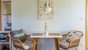Ein gemütliches Zimmer mit einem Tisch, zwei Korbstühlen, einer Wandleuchte und einem Bilderrahmen mit einem Vogelbild.