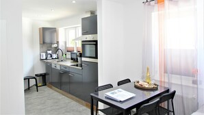 Ett modernt kök med mörka skåp, ett matbord med stolar och ett fönster med tunna, genomskinliga gardiner.