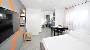 Ett modernt hotellrum med en säng, ett skrivbord, en stol, en TV, en liten köksdel och ett badrum.