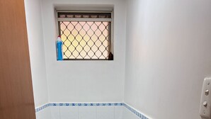 A bathroom with a toilet, blue-tiled floor, a window with a metal grid, and a door with a brass handle. - Room-cozy Room Near Airport (Perth)