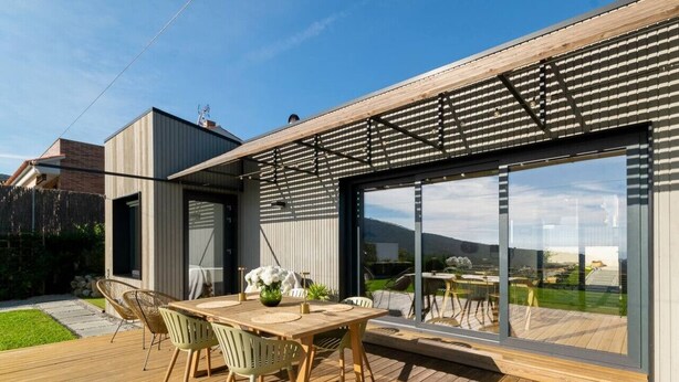 A modern house with a wooden deck, a dining area with chairs and a table, and large glass windows.