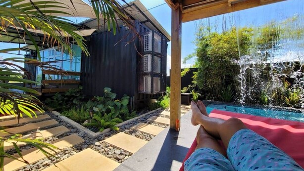 Uma pessoa relaxando numa espreguiçadeira perto de uma piscina com uma cascata, rodeada por um jardim e um edifício moderno.