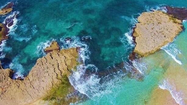 Eine Vogelperspektive eines Strandes mit klarem türkisfarbenem Wasser, Felsformationen und einer Sandküste.