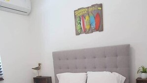 Ein ordentlich bezogenes Bett mit weißer Bettwäsche und zusammengerollten Handtüchern, ein grauer möbelbezogener Kopfteil, eine wandmontierte Klimaanlage, ein kleiner Nachttisch mit Lampe und ein Poster an der Wand.