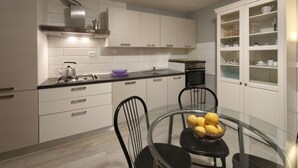 Une cuisine moderne avec des meubles blancs, une cuisinière à gaz, une hotte en acier inoxydable, une table à manger en verre avec des chaises, et un meuble encastré avec des portes en verre.