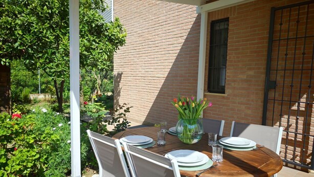 Une terrasse extérieure avec une table en bois disposée pour quatre personnes, entourée de chaises blanches, d’un vase en verre contenant des fleurs et d’un mur de briques en arrière-plan.