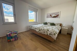 Ein Schlafzimmer mit Bett, zwei Fenstern, einem Nachttisch, einer Lampe, einem farbenfrohen Kasten und einem Hängemattengemälde.