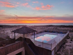 Eine Holzterrasse mit Pool und Liegestühlen, mit Blick auf einen Strand bei Sonnenuntergang.