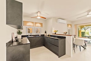 A modern kitchen with dark cabinetry, a central island, and a dining area with a table and chairs. - Marina Apartment A2, Jan Thiel (Jan Thiel)