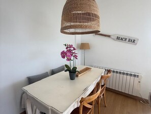 Ein Essbereich mit Tisch, Stühlen, einer Korbtaschelampe, einer Topf-Orchidee, einer Lampe und einem Schild mit der Aufschrift 'BEACH BAR'.