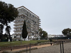 Ein mehrstöckiges Wohngebäude mit Balkonen, eine Parkanlage mit einem Weg und ein hoher Baum.