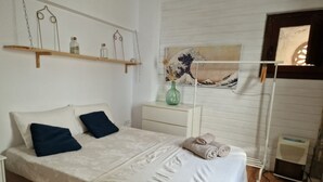 A bedroom with a bed, pillows, a bedside table, a wall-mounted shelf, a painting, and a radiator.