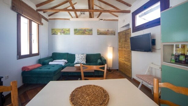 A room with a high wooden ceiling, a green sofa, a white table with a woven placemat, and a television on the wall.