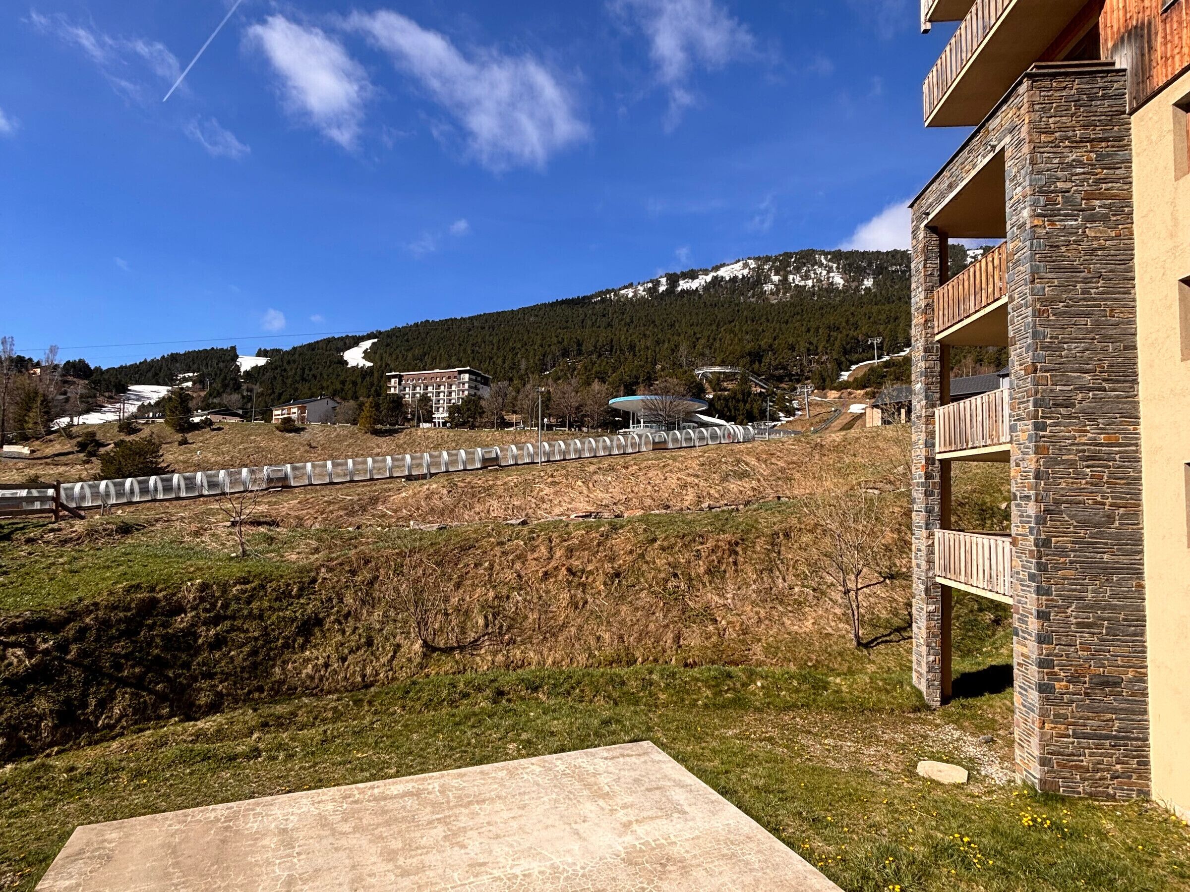 Un edificio residencial con balcones de fachada de piedra, un patio de concreto y vistas a un paisaje montañoso con una pista de esquí al fondo.