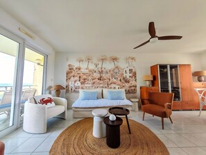 A living room with a sofa, a chair, a round rug, a ceiling fan, and a wall mural. - Bright apartment sea view - 6 guests (Canet-en-Roussillon)