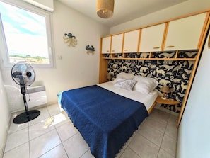 A bedroom with a bed, a standing fan, a window, and a wall with animal prints. - Bright apartment sea view - 6 guests (Canet-en-Roussillon)