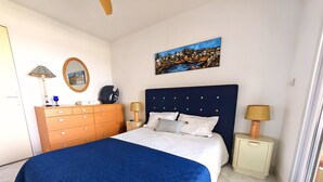 Une chambre à coucher avec un lit, une commode, une table de chevet, une lampe, un tableau et une fenêtre.