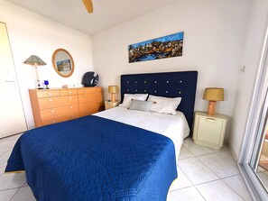 Une chambre à coucher avec un lit, une commode, une table de chevet, une lampe, un tableau et une fenêtre.
