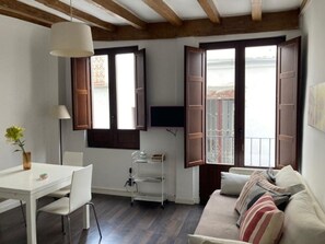 Ein Zimmer mit einem weißen Tisch, einer Couch mit gestreiften Kissen, einem an der Wand montierten Fernseher und einem Fenster mit geöffneten Fensterläden.