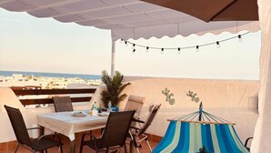 Eine Terrasse im Freien mit Tisch und Stühlen, einer Hängematte, Lichterketten und Blick auf das Meer.