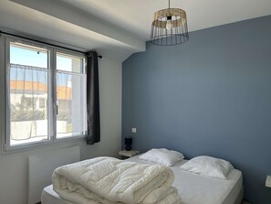 Ein Schlafzimmer mit Bett, Fenster mit Vorhängen, Nachttisch mit Lampe und einer Pendelleuchte.