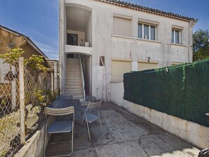 A two-story building with a staircase leading to an upper floor, a fenced outdoor area with metal chairs, and a green hedge. - Air-conditioned apartment near beach - 4 guests (Le Grau d'Agde)