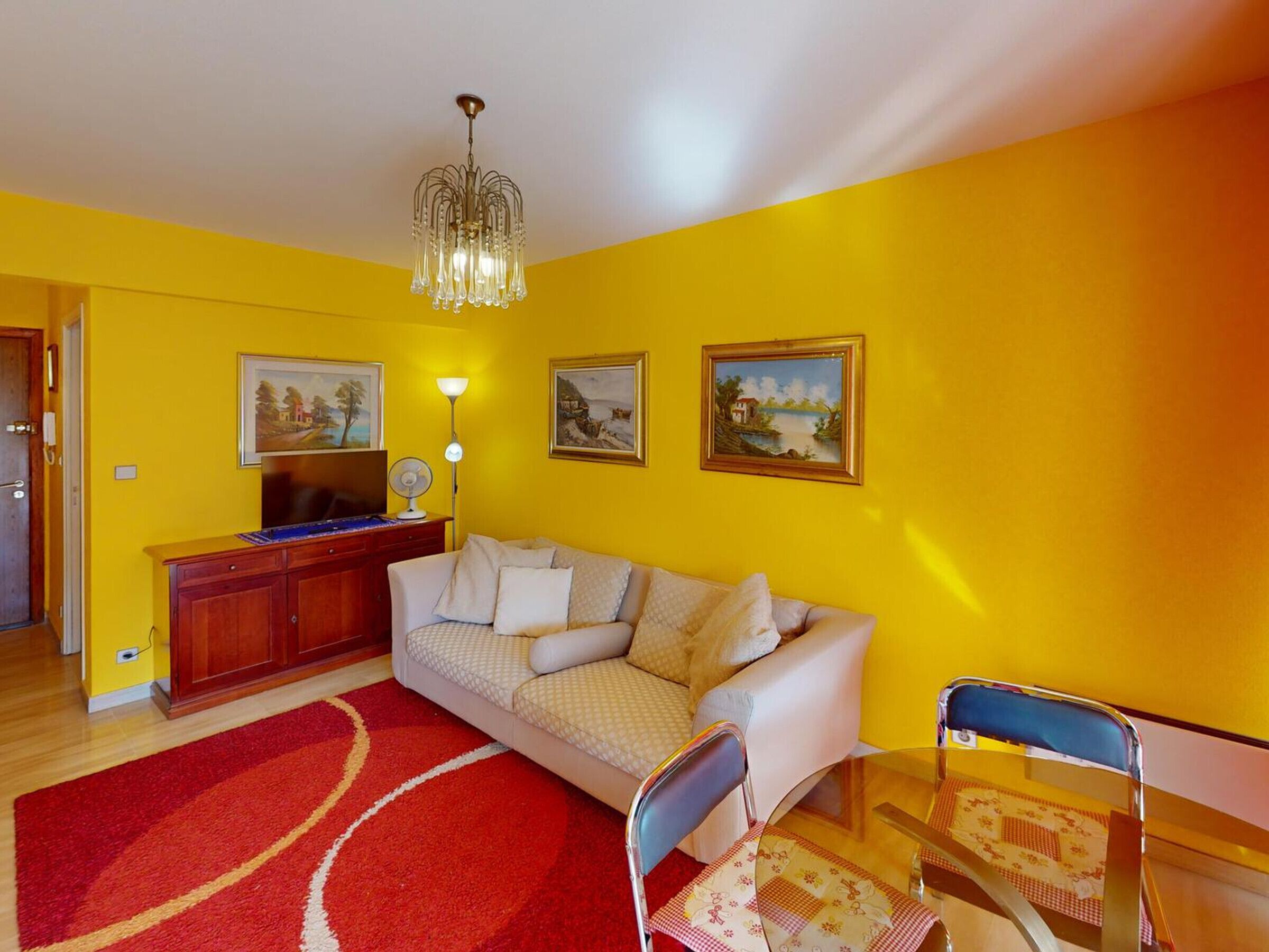 A living room with a sofa, a TV, a chandelier, and paintings on the wall.