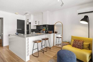 A modern kitchen with a central island, bar stools, a yellow sofa, and a large mirror. - Galleries Apartments, 1 Bed, Parking, Balcony (Fortitude Valley)