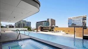 Una piscina en la azotea con vistas a la ciudad, cielo despejado y edificios modernos al fondo.