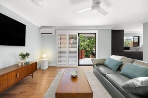 A modern living room with a sofa, coffee table, TV, and a sliding glass door leading to a balcony. - Spacious 2BR Stay w/ Pool & Balcony (Auchenflower)