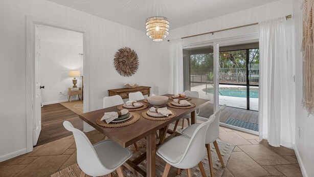 Un comedor con mesa de madera, sillas blancas, una lámpara de araña y vista a una piscina.