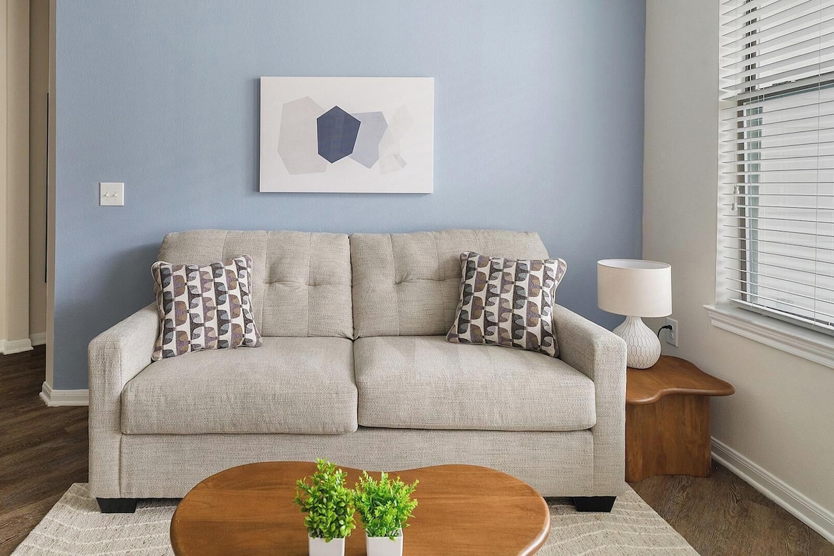 A living room with a sofa, two patterned pillows, a wall-mounted abstract art piece, a side table with a lamp, and a small wooden coffee table with plants.