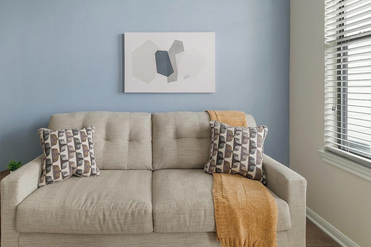 A beige sofa with patterned cushions, a white wall art piece, a wooden side table with a lamp, and a window with blinds.