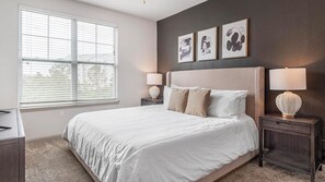 A bedroom with a large bed, two bedside tables, a window with blinds, and three wall-mounted artworks. - Landing Apartments near Gateway Crossing (McCordsville)