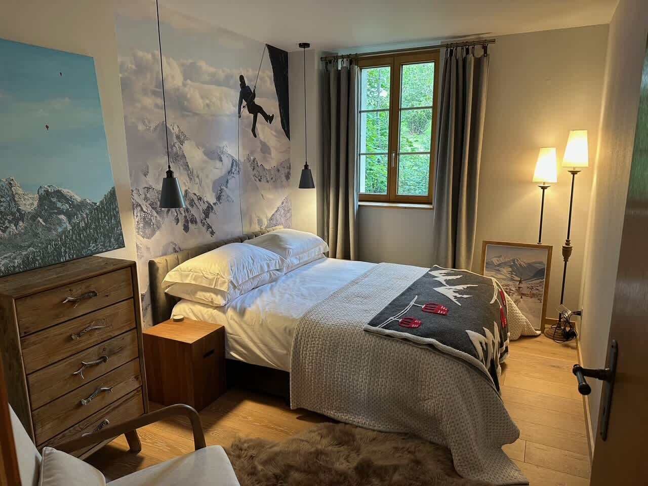 A bedroom with a bed, a dresser, a nightstand, a window with curtains, and a wall-mounted lamp.