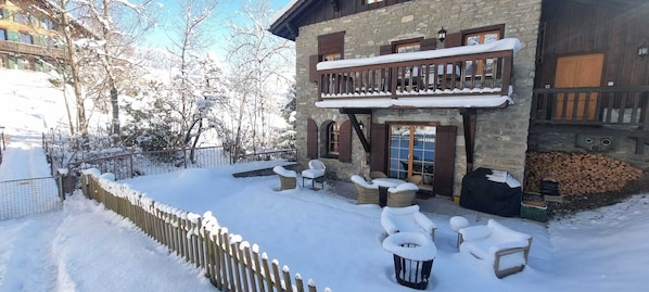 Un espace extérieur aménagé avec des sièges, recouvert de neige, comprenant des chaises en osier et un bâtiment en pierre doté d’un balcon en bois.