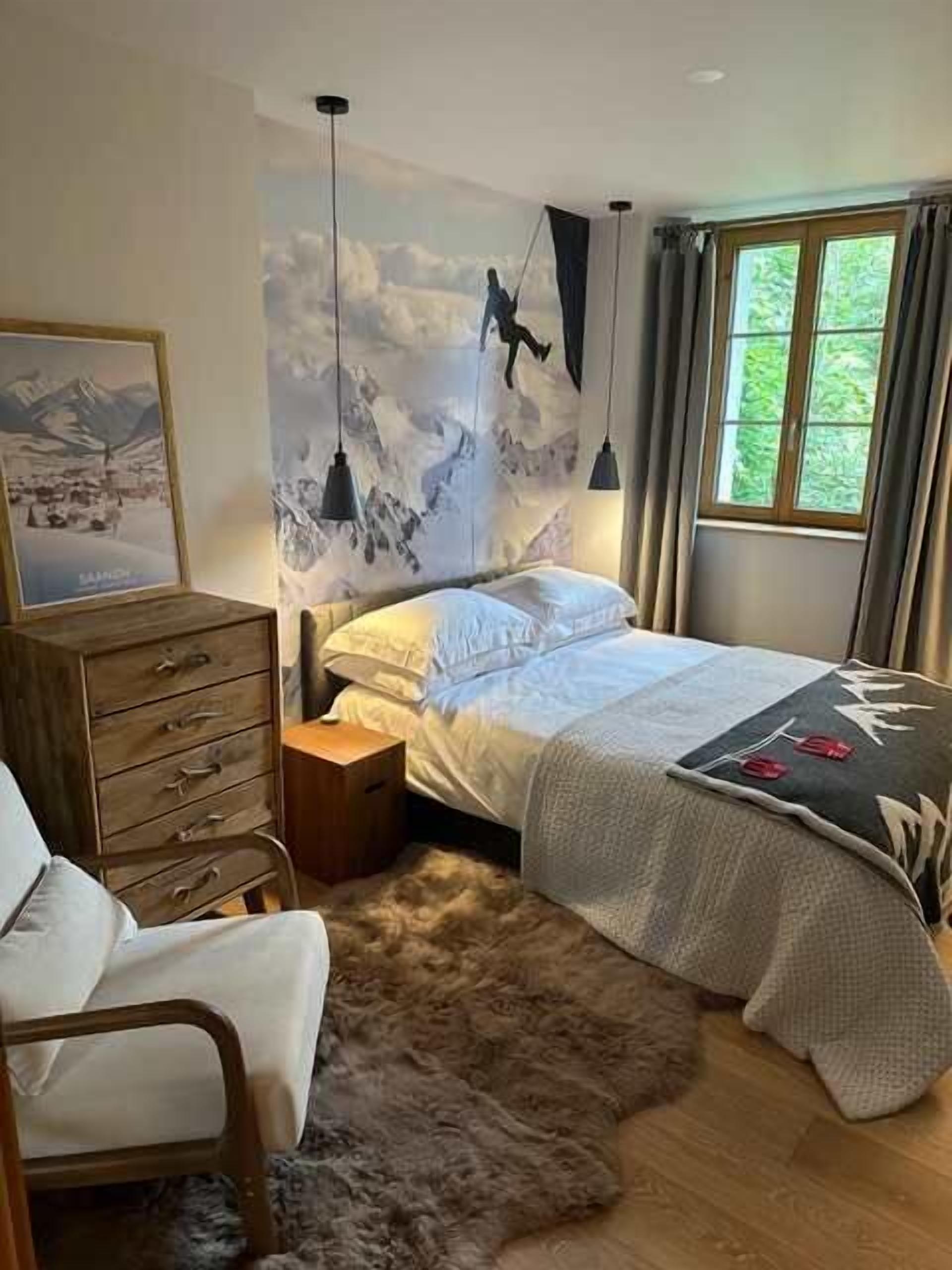 Une chambre à coucher avec un lit, une commode en bois, une chaise, une table de chevet, un mur orné d’un tableau représentant une montagne et une fenêtre avec des rideaux.