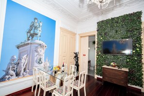 Ein Essbereich mit einem Glastisch, weißen Stühlen, einem großen Wandgemälde, einem Fernseher und einer begrünten Wand.