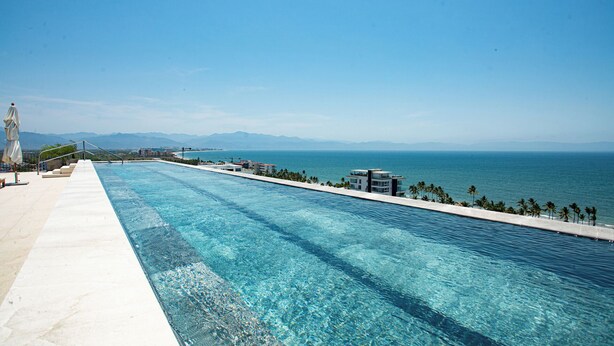 A long swimming pool with clear blue water, a white border, and a view of the ocean and distant mountains. - Oceana Luxury Escape | 3BR/3BA | Ocean Views (Bucerias)
